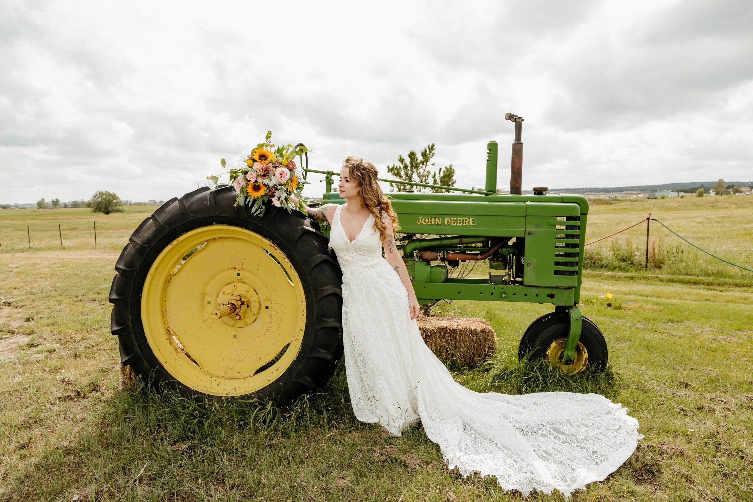Bride in white wedding dress beside vintage tractor in rural farmland